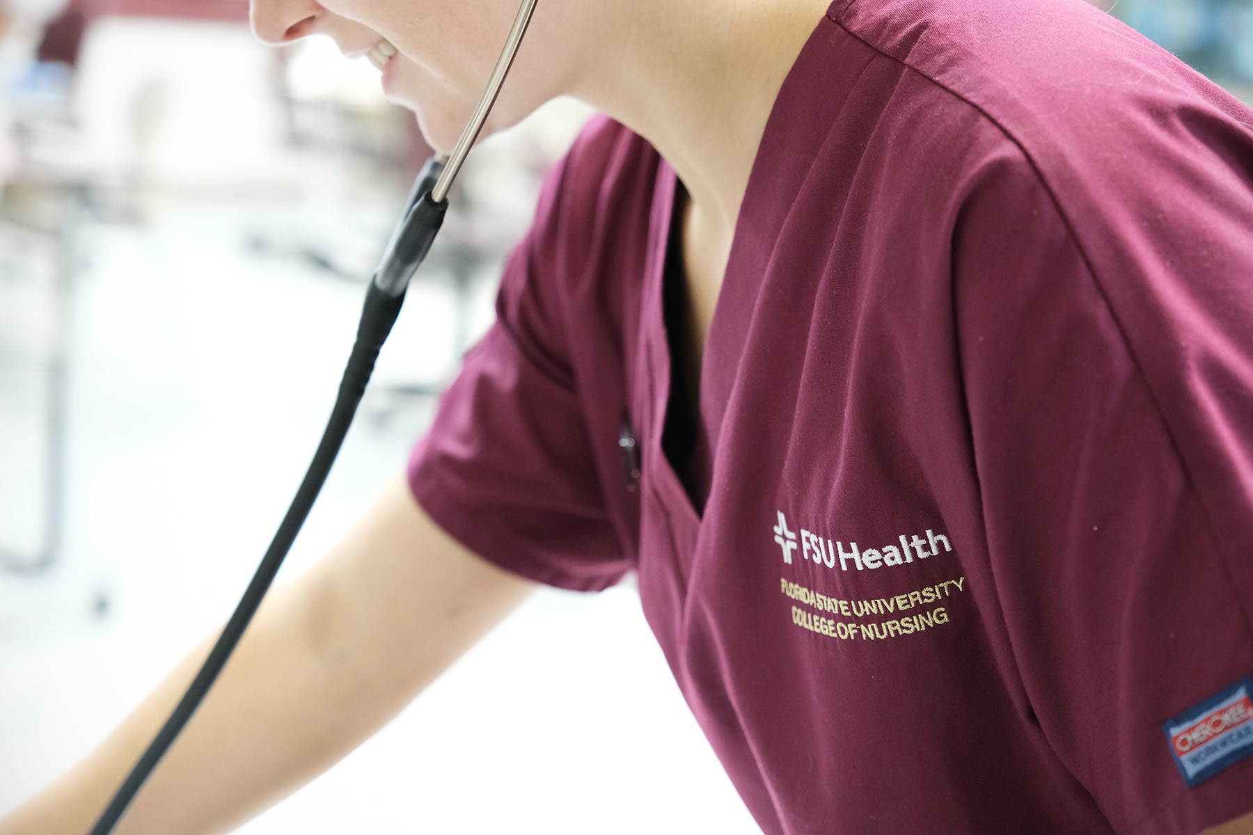 A photo of a nursing student wearing Garnet scrubs, smiling and using a stethoscope