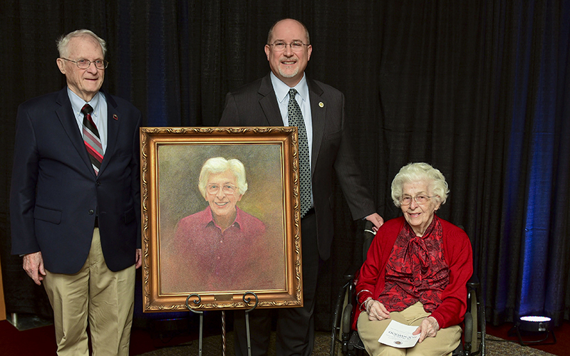 Photo of 2 men and a woman around a painted portrait of the woman