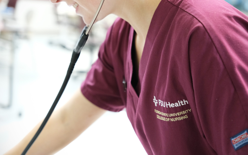 A photo of a nursing student wearing Garnet scrubs, smiling and using a stethoscope